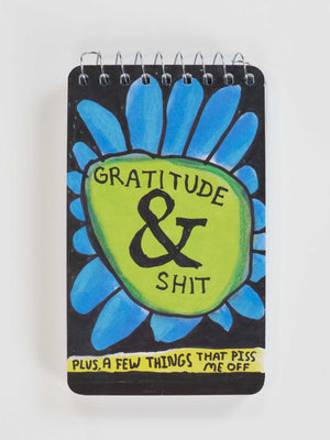 The Blue Q “Gratitude & Shit. Plus A Few Things That Piss Me Off” 3x5 spiral notebook features blue petals, a green center, and a yellow band—perfect for tracking gratitude or jotting down daily annoyances.