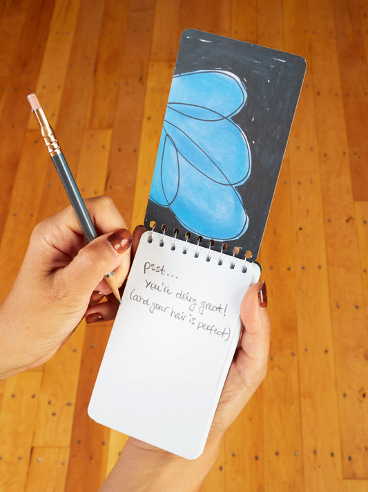 A person holds the Blue Q "Gratitude & Shit. Plus A Few Things That Piss Me Off" 3x5 Spiral Notebook and a pencil. The notepad has a handwritten note: psst... you're doing great! (and your hair is perfect), with a blue flower above.