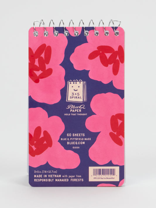 Introducing the Blue Q “You’re Beautiful Don’t Change” 3x5 Spiral Notebook—a compact notebook featuring bold pink and red flowers on a navy background. Perfect as a motivational gift for the amazing weirdo in your life, it includes branding, website, eco-friendly details, and a “You’re Beautiful” sticker on the back cover.