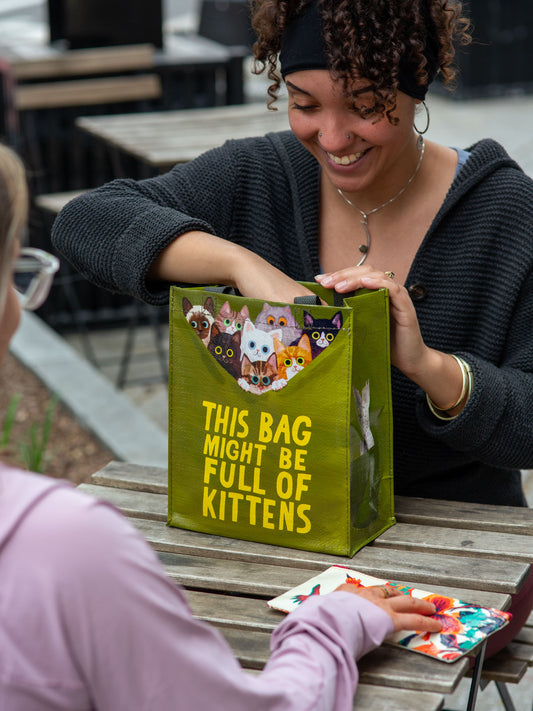 A smiling woman at an outdoor table reaches into a green Blue Q "This Bag Might Be Full Of Kittens Handy Tote"—the playful design matches the scene as adorable kittens peek out. Another person sits across from her.