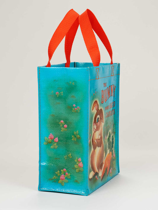 The Bunny Who Hated Carrots Handy Tote by Blue Q features a turquoise design with red handles, a whimsical bunny and flowers, and is perfect for carrying your favorite book on the go.