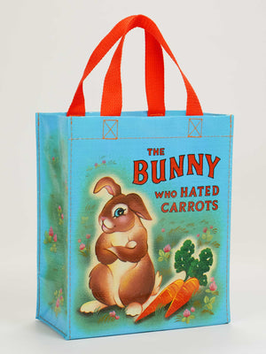 The Bunny Who Hated Carrots Handy Tote by Blue Q is a blue bag with red handles, featuring a bunny illustration with two carrots and green plants, plus the title text for a whimsical storybook vibe.