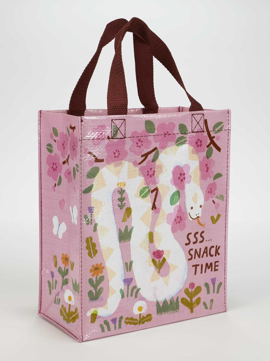 The Blue Q Sss...Snack Time Handy Tote is a pink reusable bag with brown handles, eco-friendly materials, and a white snake among pink flowers and greenery. "SSS...SNACK TIME" is printed beside the design.