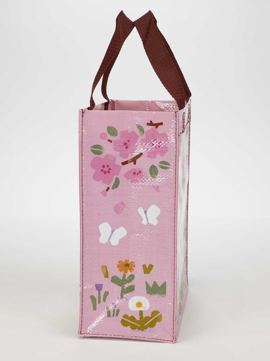 The Sss...Snack Time Handy Tote by Blue Q is a pink, eco-friendly reusable bag with brown handles, featuring vibrant flowers, butterflies, and leaves—ideal for snacks or daily errands.
