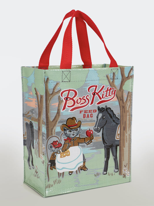 The Blue Q Boss Kitty Handy Tote is a reusable bag with red handles, featuring a cat in a cowboy hat feeding an apple to a black horse in a forest. The text "Boss Kitty Feed Bag" makes this eco-friendly tote fun and practical.
