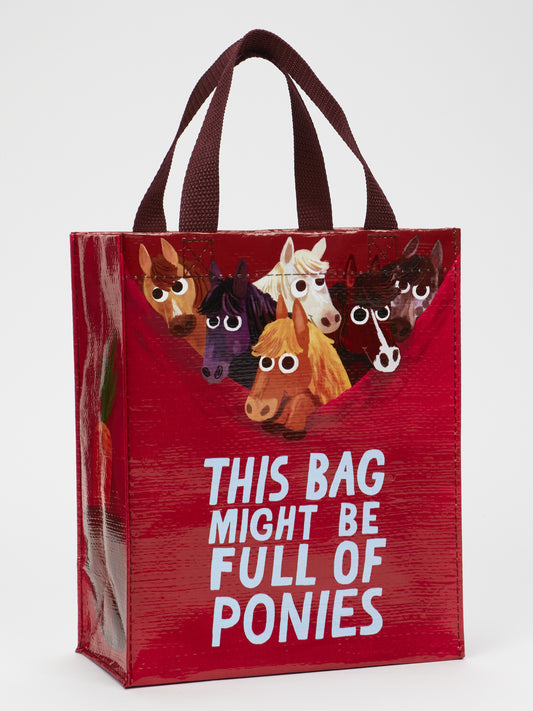 The Blue Q "This Bag Might Be Full Of Ponies Handy Tote" is a red eco-friendly bag with maroon handles, cartoon ponies peeking over the top, and "This bag might be full of ponies" in bold white letters on the front.
