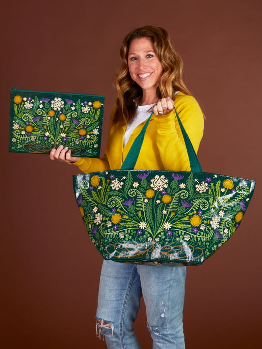 A smiling woman in a yellow cardigan and jeans holds a green floral tote made from recycled material and Blue Q's Wild Bloom Jumbo Pouch, both featuring botanical designs, set against a brown background.