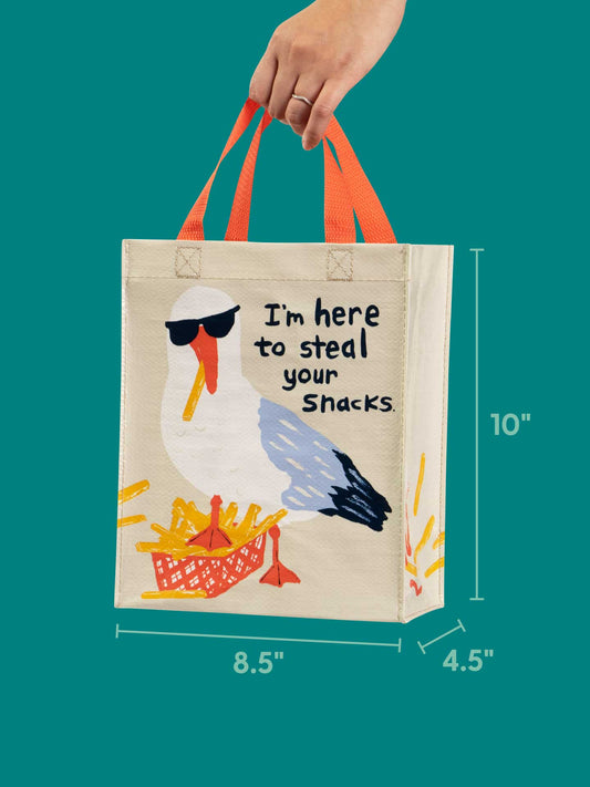 A hand holds the Blue Q "I'm Here To Steal Your Snacks Handy Tote," featuring orange handles and a seagull in sunglasses next to fries with the text "I'm here to steal your snacks." Bag size: 10 x 8.5 x 4.5 inches.