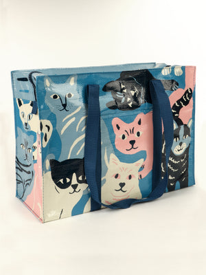 The Blue Q Happy Cats Shoulder Tote is a reusable bag featuring illustrated cats in blue, pink, cream, and black. It has thick blue handles, a glossy finish, and is shown on a plain white background.