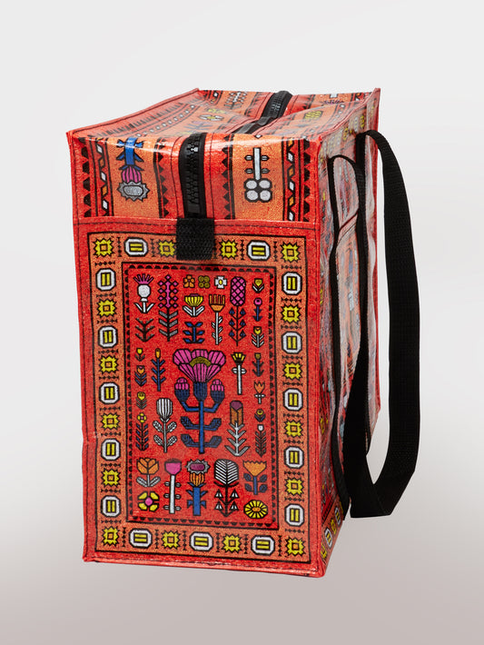 The Magic Carpet Shoulder Tote by Blue Q is a durable bag made from recycled material with black handles, a secure zipper, and an eye-catching mix of red, yellow, blue, and white geometric and floral patterns on a textured surface.