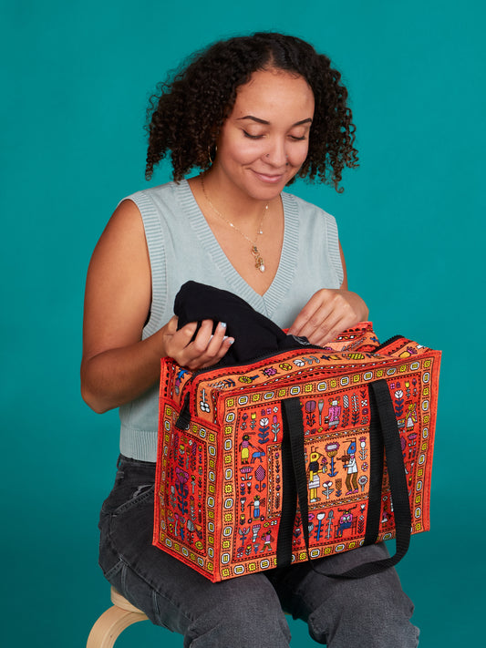 A woman with curly hair wearing a light blue sleeveless top and jeans smiles as she places a black item into the Blue Q Magic Carpet Shoulder Tote, featuring a secure zipper, against a teal background.