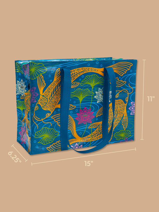 The Blue Q Koi Shoulder Tote is a reusable shopping bag featuring blue handles, crafted from recycled material and adorned with cheerful orange koi fish, lotus flowers, and green lily pads on a tan background. The bag measures 15" wide, 11" tall, and 6.25" deep.