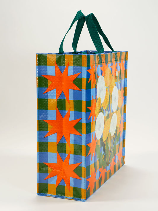 The Blue Q Dandelion Pop Shopper is a colorful reusable bag with green handles, featuring a blue and yellow checkered pattern, orange stars, and white dandelion flower illustrations.