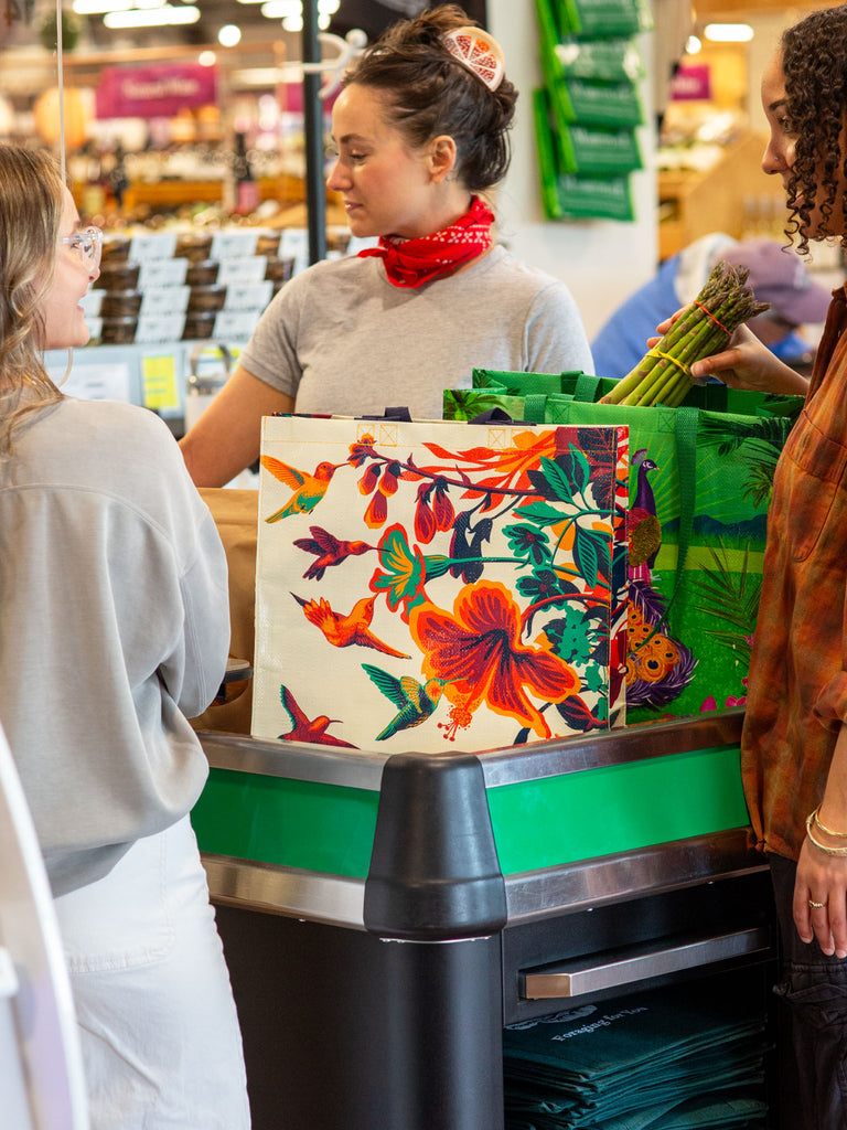 Three women check out at a grocery store with vibrant reusable bags, including the Blue Q Hummingbird Shopper, which features colorful birds and flowers. One woman holds a bunch of asparagus.