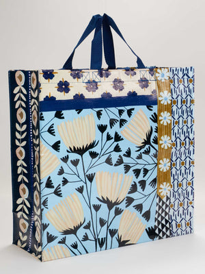 The Blue Q Patchwork Shopper is a spacious bag with blue handles, featuring geometric patterns and floral designs in blue, cream, black, and gold on a light blue background—perfect for shopping or storage.
