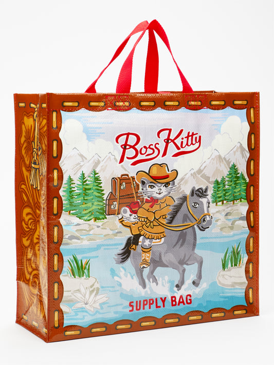 The Blue Q Boss Kitty Shopper is a reusable grocery bag featuring a cartoon cat dressed as a cowboy riding a horse across a river, with mountains and trees in the background. The text on the eco-friendly shopping bag reads "Boss Kitty Supply Bag," and it comes with sturdy red handles.