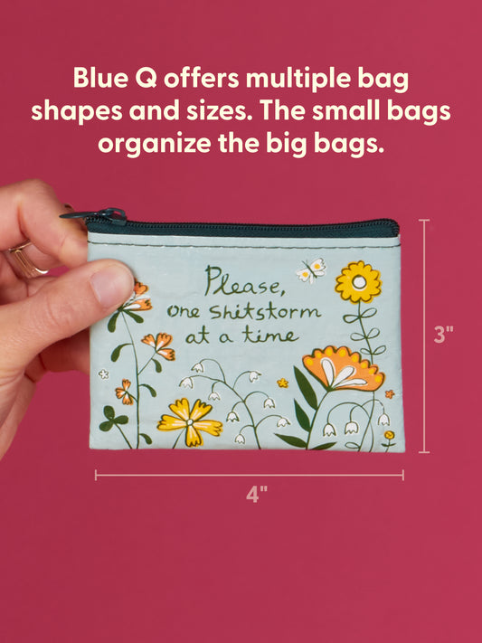 A hand holds the Blue Q "Please, One Shitstorm At A Time" Coin Purse, crafted from recycled material with colorful flowers on a red background. Measuring 3"x4", it’s a fun, eco-friendly gift option.