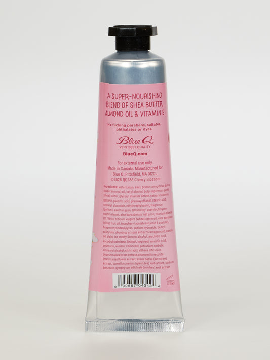 A pink and silver tube of Blue Q's "I'm A Delicate Fucking Flower" hand cream stands upright, featuring wild cherry blossom with sandalwood, plus shea butter and almond oil, along with product details and instructions on the back.