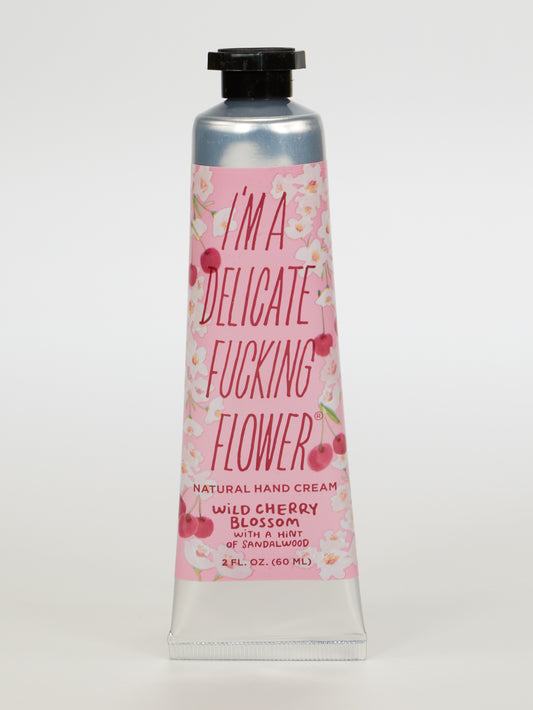 Blue Q’s I'm A Delicate Fucking Flower Natural Hand Cream—Wild Cherry Blossom with a hint of Sandalwood—comes in a 2 fl. oz. pink tube with bold text and floral designs, blending shea butter and almond oil for soft, nourished hands.