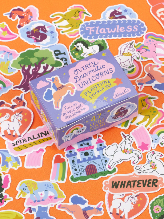 A purple Blue Q Overly Dramatic Unicorns Playtime Sticker Set box surrounded by various unicorn-themed stickers on an orange surface.