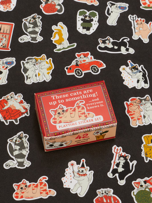 A red box labeled “These Cats Are Up To Something! Playtime Sticker Set” by Blue Q rests on black fabric, surrounded by playful, colorful cat stickers in various costumes and poses—an adorable gift idea that’s perfect for anyone who loves novelty figurines or is searching for fun gifts for teens.