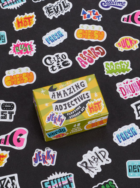 A yellow box labeled Amazing Adjectives Playtime Sticker Set from Blue Q is surrounded by a vibrant array of adjective stickers featuring descriptive words like jerky, hot, rude, soggy, and more—all designed to help modify nouns—set against a black background.