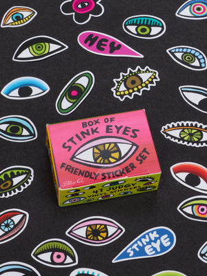 A vibrant Blue Q Box Of Stink Eyes Friendly Sticker Set rests on a surface scattered with diverse, illustrated eye stickers in assorted colors and styles—making it the ideal desk accessory or quirky gift for friends who appreciate a bit of playful flair.