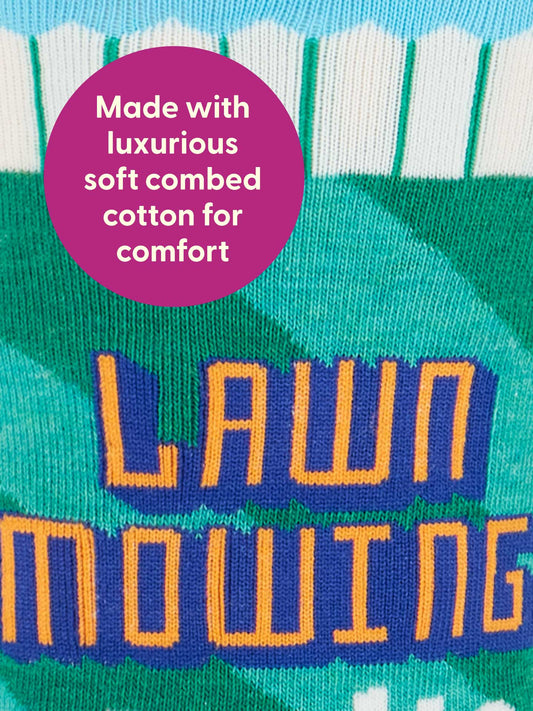 A close-up of Blue Q's Lawn Mowing Socks M-Crew Socks shows "LAWN MOWING" in orange and purple with a white fence and green grass design. A purple circle notes: Made with luxurious soft combed cotton for comfort.