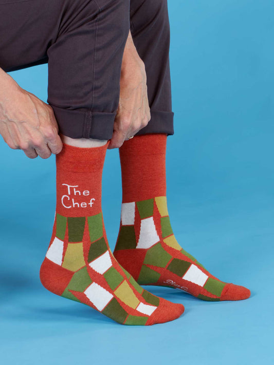 A man sits against a blue background, adjusting his pants to reveal Blue Q's "The Chef" Men's Socks—combed cotton socks in red, green, and white geometric patterns, fitting men's shoe sizes 7–12. One sock features the text "The Chef.