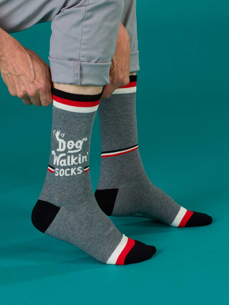 A person adjusts their Blue Q Dog Walkin' Men's Socks with paw prints and "Dog Walkin'" text. The cotton socks, in black, red, and white, are worn over gray pants against a teal background.
