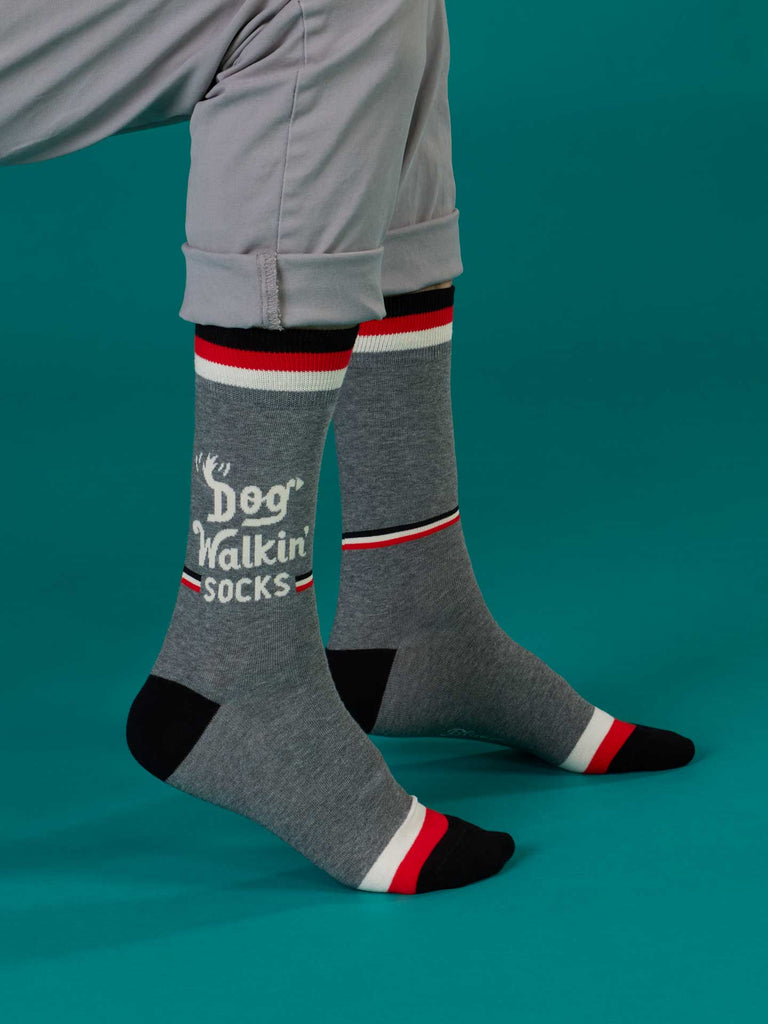 A person wears Blue Q Dog Walkin' Men's Socks—gray cotton socks with "Dog Walkin' Socks" in white, featuring black, white, and red stripes at the top, toes, and heels—styled with light gray rolled pants on a teal background.