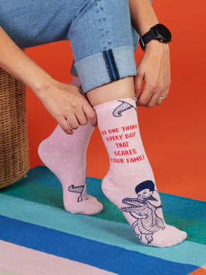 A person in rolled-up jeans puts on Blue Q’s Do One Thing Every Day That Scares Your Family W-Crew Socks—pink combed cotton socks showing a child wrestling an alligator, red text, and an orange background atop a multicolored rug.