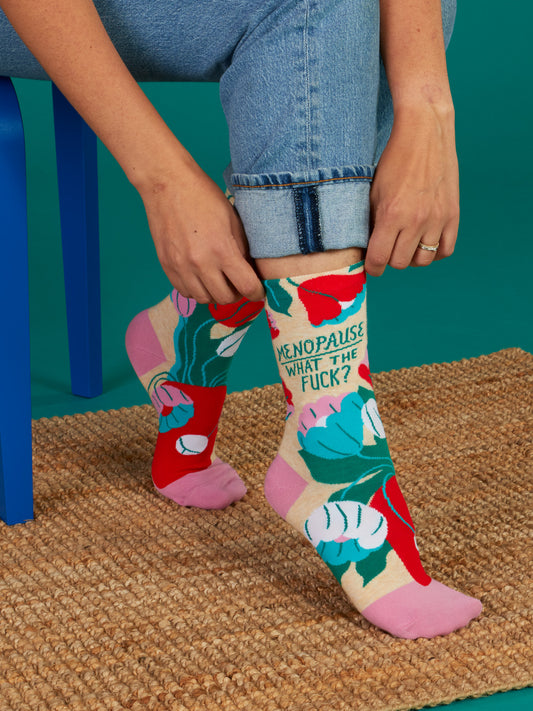 A person in rolled-up jeans sits on a blue chair, adjusting Blue Q’s "Menopause. What The Fuck? W-Crew Socks," featuring colorful floral patterns, while on a woven mat against a teal background.