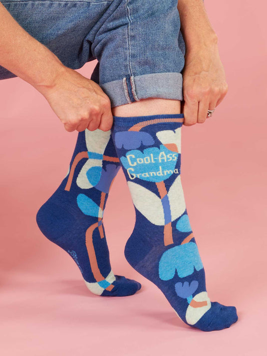 A person wearing Blue Q Cool-Ass Grandma Crew Socks—women's size 5-10, with colorful patterns and "Cool Ass Grandma" text—adjusts their rolled-up jeans against a pink background.