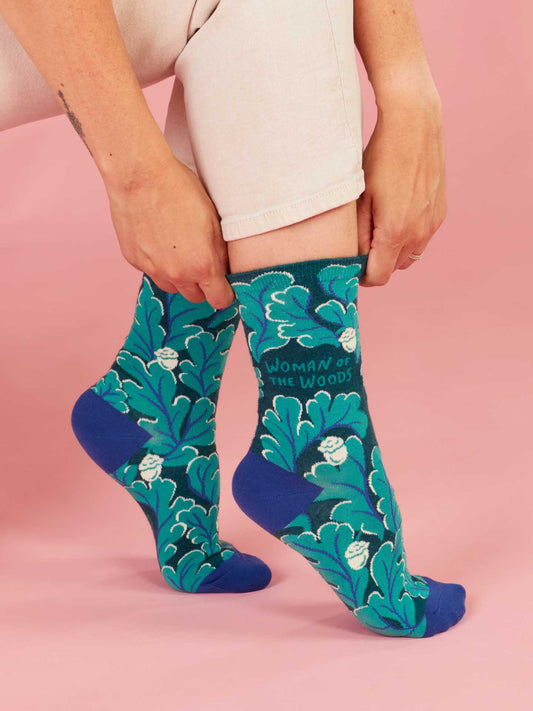 A person adjusts one Blue Q Woman Of The Woods Crew Sock—featuring green and blue leafy patterns, berry print, and "WOMAN OF THE WOODS" text—while sitting on a pink background. Only their lower legs and hands are visible.
