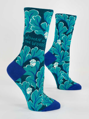 Blue Q Woman Of The Woods Crew Socks for women feature blue-green cotton with leafy patterns, small white faces, foraged berry accents, blue heels and toes, and "WOMAN OF THE WOODS" in teal on the side.