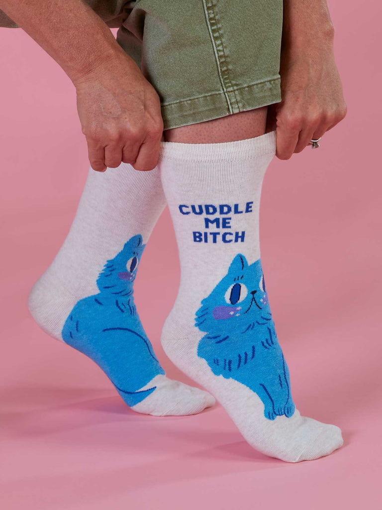 A person wears Blue Q's Cuddle Me Bitch Crew Socks featuring a blue cartoon cat and bold text, standing on a pink background.