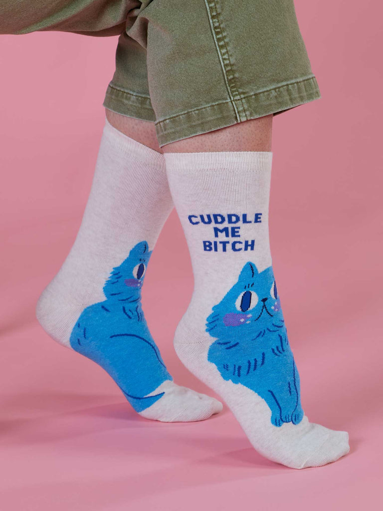 A person wearing Blue Q's Cuddle Me Bitch Crew Socks—light-colored women’s socks with a bold blue cat design and “CUDDLE ME BITCH” text—stands on a pink background, paired with green pants.