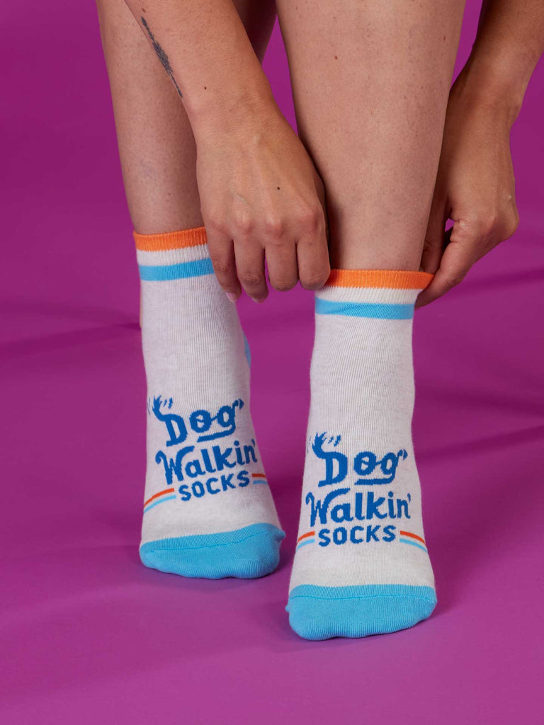 A person on a purple background adjusts a pair of Blue Q Dog Walkin' Ankle Socks, featuring blue toes, blue accents, orange trim, and "Dog Walkin’ Socks" in blue on the front.