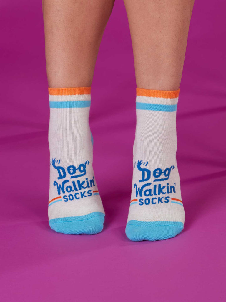 A person wears Blue Q Dog Walkin' Ankle Socks—white with blue toes and heels, orange and blue stripes, and "Dog Walkin socks" text—while standing on a purple surface. Perfect for daily dog walks.