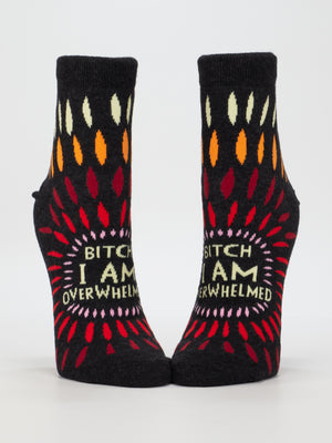 The Blue Q Bitch I AM Overwhelmed Ankle Socks are black combed cotton socks with colorful patterns and bold lettering on the soles, designed to fit women’s shoe size 5-10.