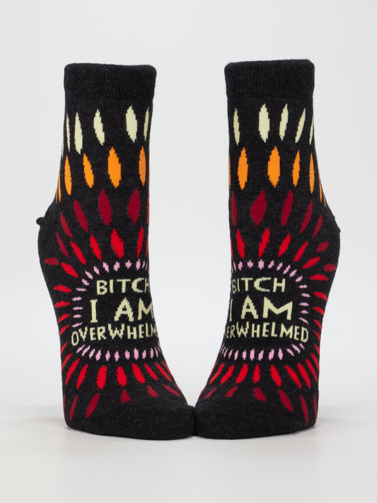 The Blue Q Bitch I AM Overwhelmed Ankle Socks are black combed cotton socks with colorful patterns and bold lettering on the soles, designed to fit women’s shoe size 5-10.