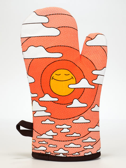 The Blue Q "Somebody Get Me A Gummy Oven Mitt" features a vibrant orange and red swirl design with a smiling yellow sun and white clouds. Made from 100% cotton, it combines cheerful style, comfort, and durable protection for your kitchen.
