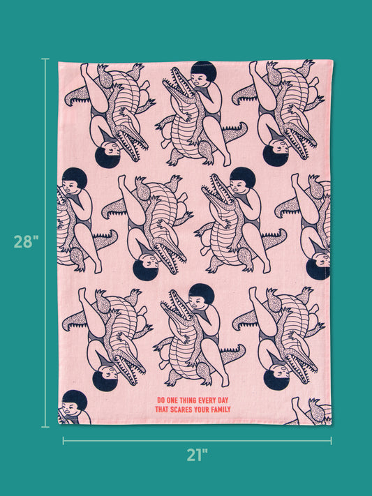 The Blue Q Do One Thing Every Day That Scares Your Family Dish Towel (21x28), pink with a child wrestling a crocodile and the phrase at the bottom, is displayed on a teal background.