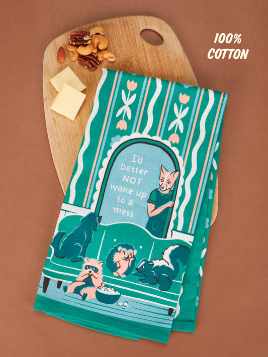 The Blue Q "I'd Better Not Wake Up To A Mess" dish towel, made of 100% cotton and featuring colorful animals, is shown on a wooden cutting board with cheese slices and nuts, set on a brown surface.