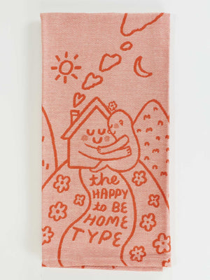 The Happy To Be Home Type Dish Towel by Blue Q features red illustrations of a smiling house, flowers, sun, moon, and hearts on pink cotton, with “The happy to be home type” text. Ideal for those who cherish solitude or calm.