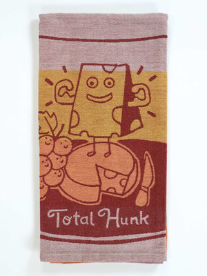 The Blue Q Total Hunk Dish Towel is super-absorbent and shows a cartoon cheese block with arms, flexing on cheese wheels beside grapes. The phrase "Total Hunk" appears at the bottom.