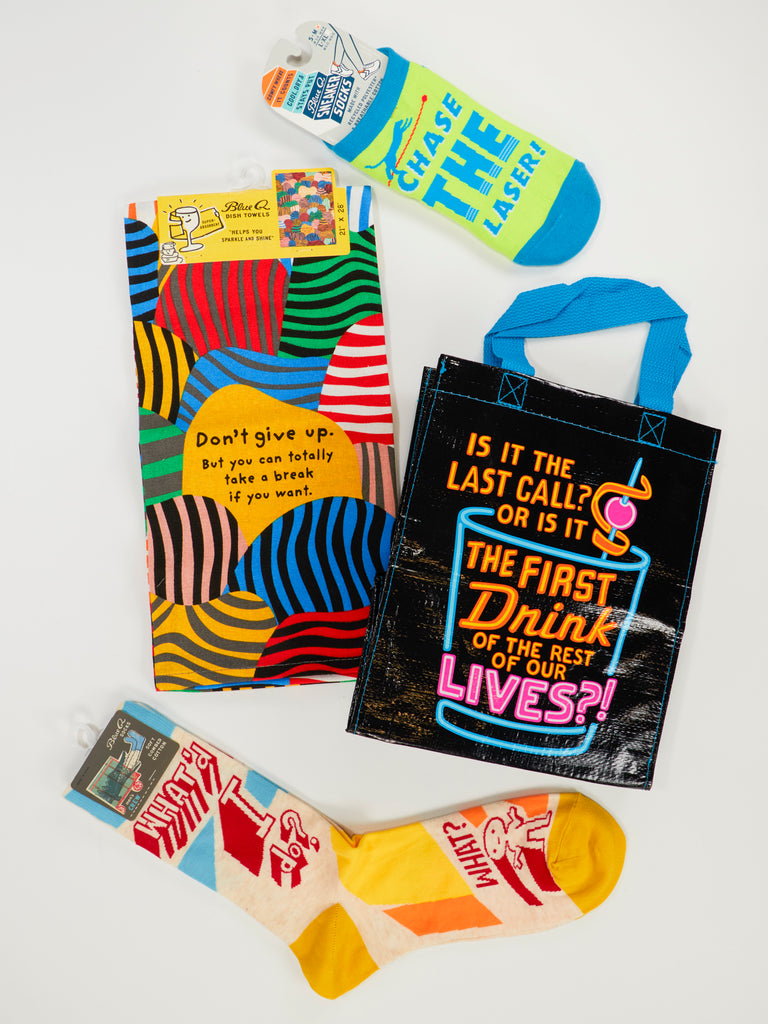 The Blue Q Fun For All Sale Pack features colorful novelty socks with motivational phrases, a quirky dish towel, and a reusable bag that says, "Is it the last call or the first drink of the rest of our lives?.